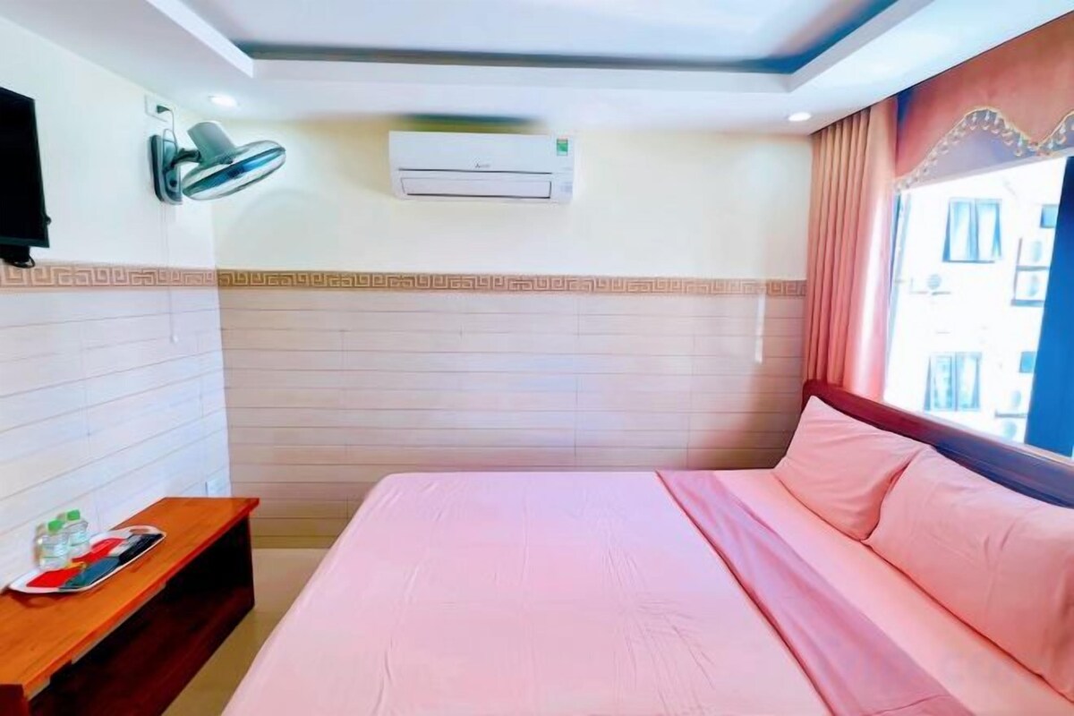 A hotel room with a bed, a bedside table, a TV, an air conditioning unit, and a window with curtains.