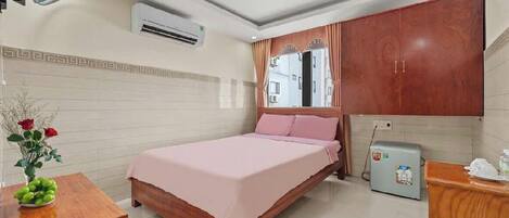 A bedroom with a bed, a wooden nightstand, a small table with a vase of flowers, an air conditioner, a ceiling fan, a wardrobe, and a refrigerator.