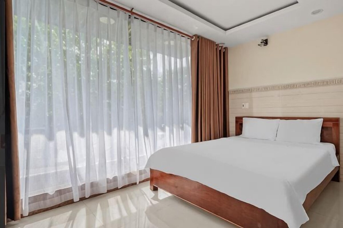 A hotel room with a large bed, a window with sheer curtains, and a view of greenery outside.