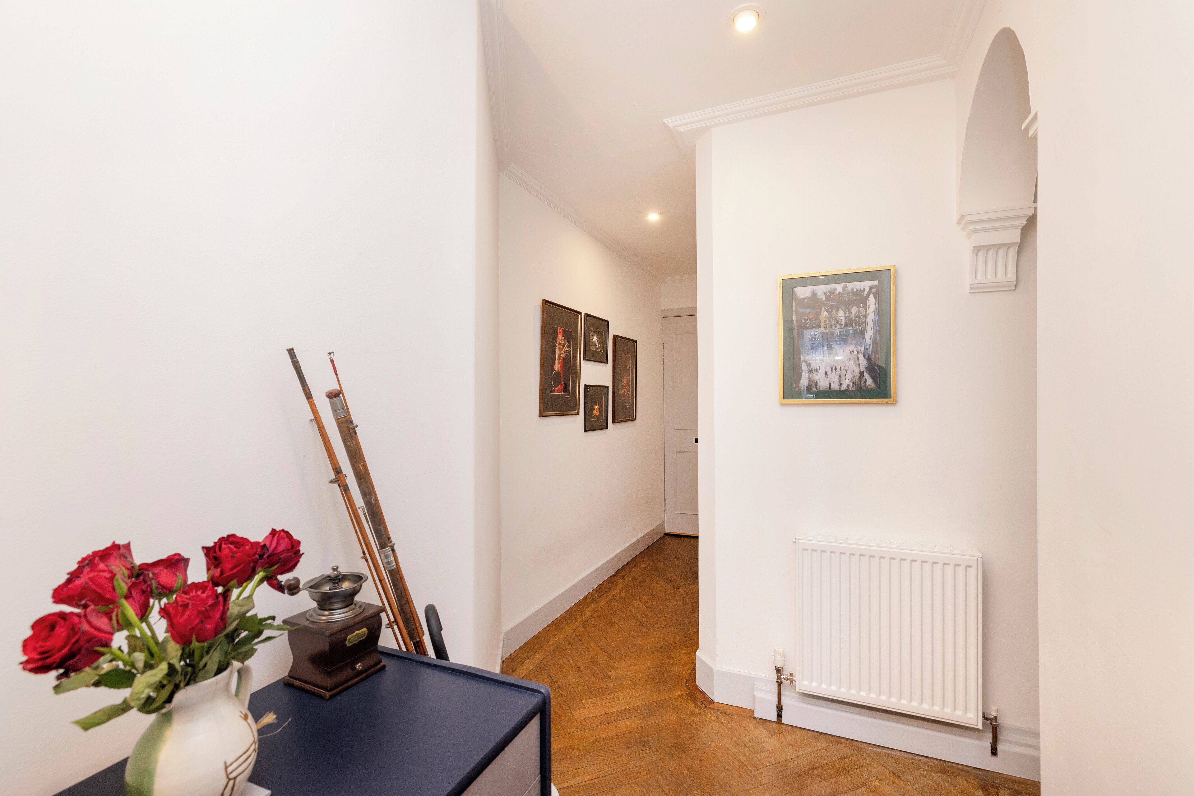 Un pasillo con piso de madera, paredes blancas y un radiador. Hay cuadros enmarcados en la pared y un florero con rosas rojas sobre una mesa.