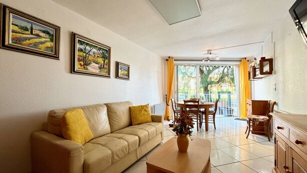 A living room with a beige sofa, a wooden coffee table, and a dining table with chairs. There are framed pictures on the wall and a window with curtains.