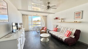 Ein geräumiger Wohnbereich mit Sofa, Couchtisch und Esstisch. Es gibt einen Deckenventilator, ein Fenster mit Vorhängen und ein gerahmtes Bild an der Wand.
