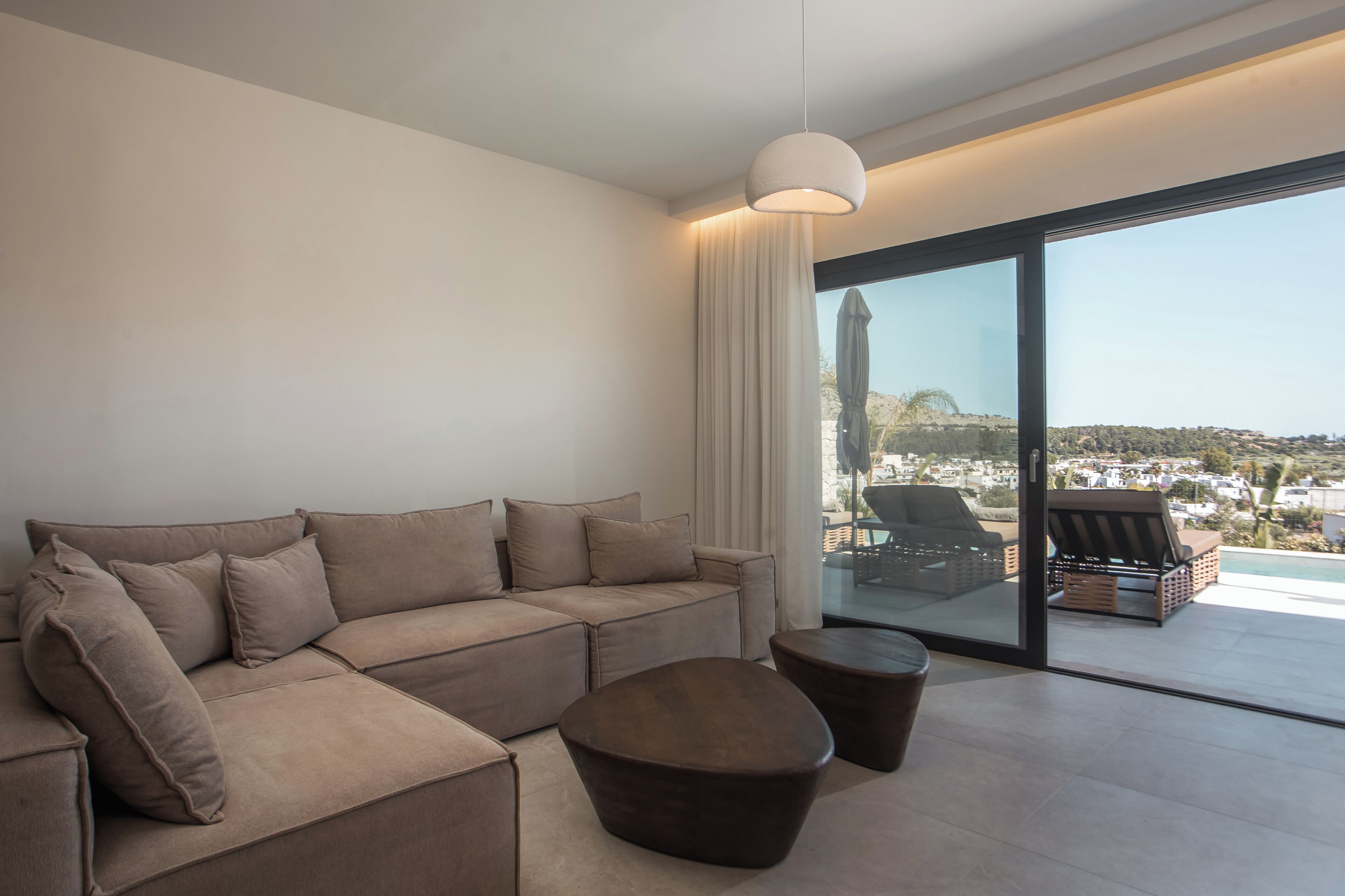 Una sala de estar moderna con un sofá seccional, una mesa de centro redonda y un gran ventanal corredizo que da a una zona de estar exterior con vistas a un paisaje urbano.