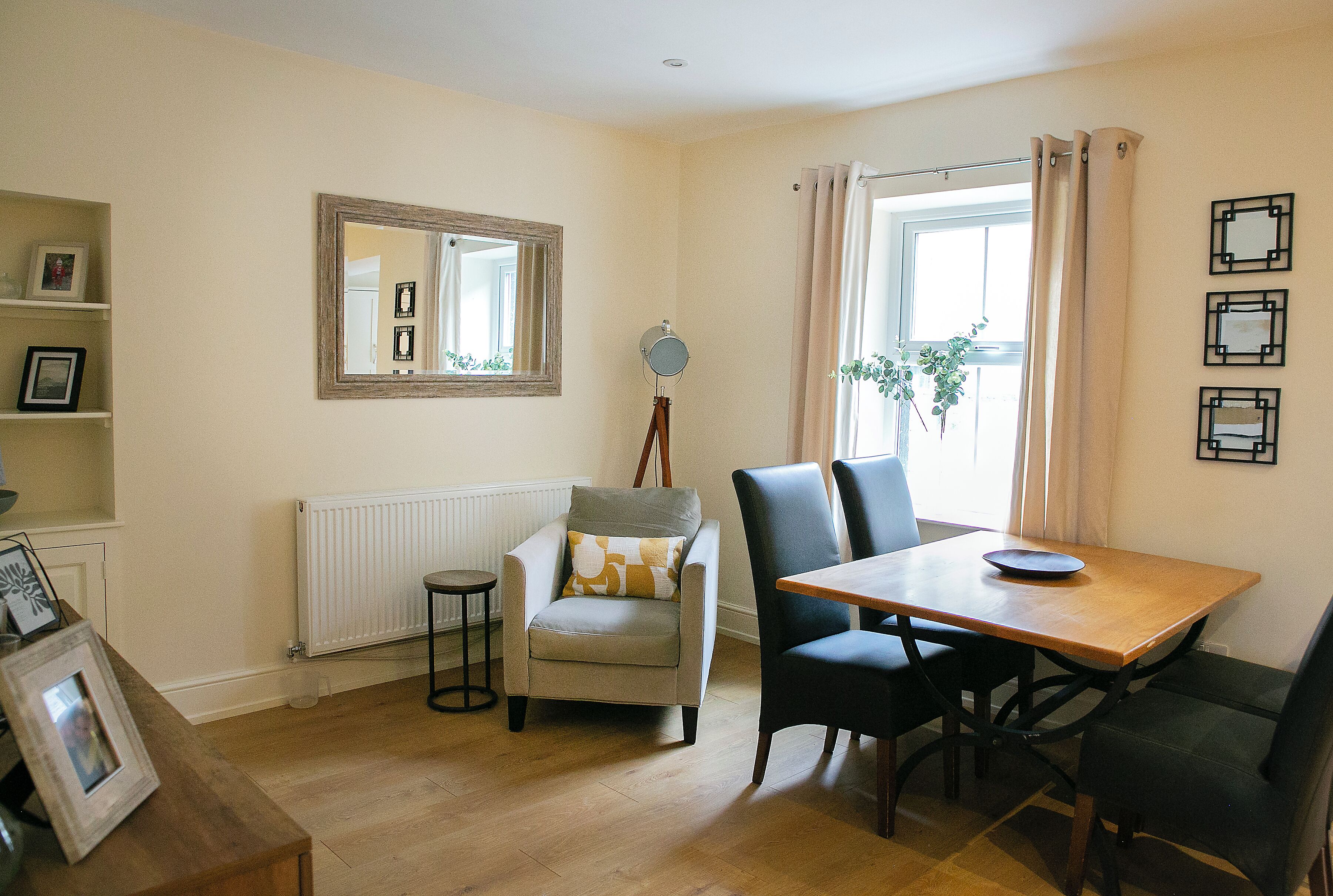 Een kamer met een houten eettafel, twee stoelen, een fauteuil, een spiegel en een radiator.