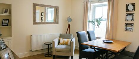 Een kamer met een houten eettafel, twee stoelen, een fauteuil, een spiegel en een radiator.