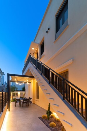 A modern building with a staircase leading to a balcony, a dining area with outdoor seating, and a well-lit entrance. - Kyma Luxury Apartment (Ρόδος)