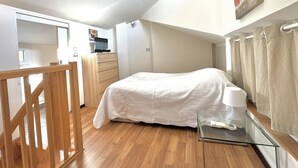 Una camera da letto con un letto, una cassettiera, una scrivania con un computer e un tavolino in vetro con una lampada.