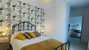 Un dormitorio con cama, mesita de noche, lámpara y pared con estampado de hojas.