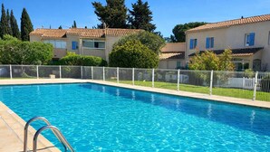 Una piscina rectangular con agua azul cristalina, rodeada por una cerca blanca y adyacente a edificios con tejados de tejas.
