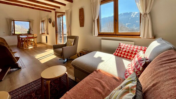 Een gezellige woonkamer met een bank, fauteuil en een eettafel. Er zijn grote ramen met gordijnen, een uitzicht op bergen en houten balken in het plafond.