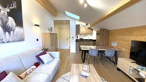 Ein modernes Wohnzimmer mit einer Couch, einem Couchtisch und einem Bild von einem Reh. Es gibt eine Küchenzeile mit Holzschränken und einen Esstisch mit Stühlen.