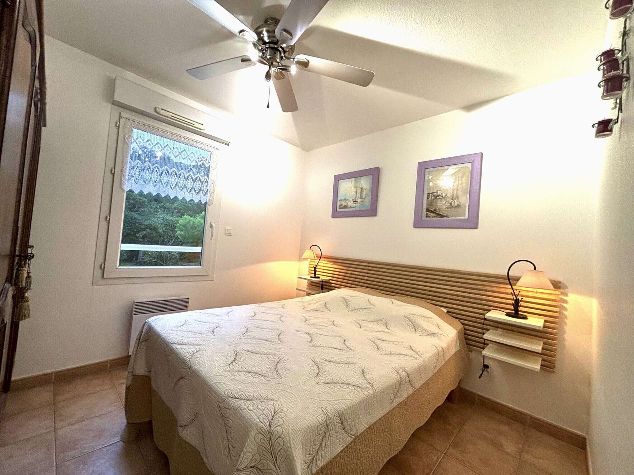 A bedroom with a bed, a ceiling fan, a window with a view, and two framed pictures on the wall.