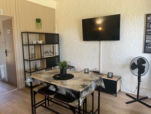 A modern living room with a TV mounted on the wall, a dining table with a tropical leaf-patterned tablecloth, a black metal bookshelf, and a standing fan.