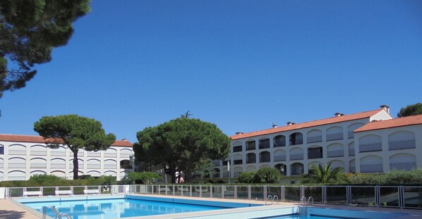 A swimming pool with a white fence, a large tree, and a building with a red roof in the background.