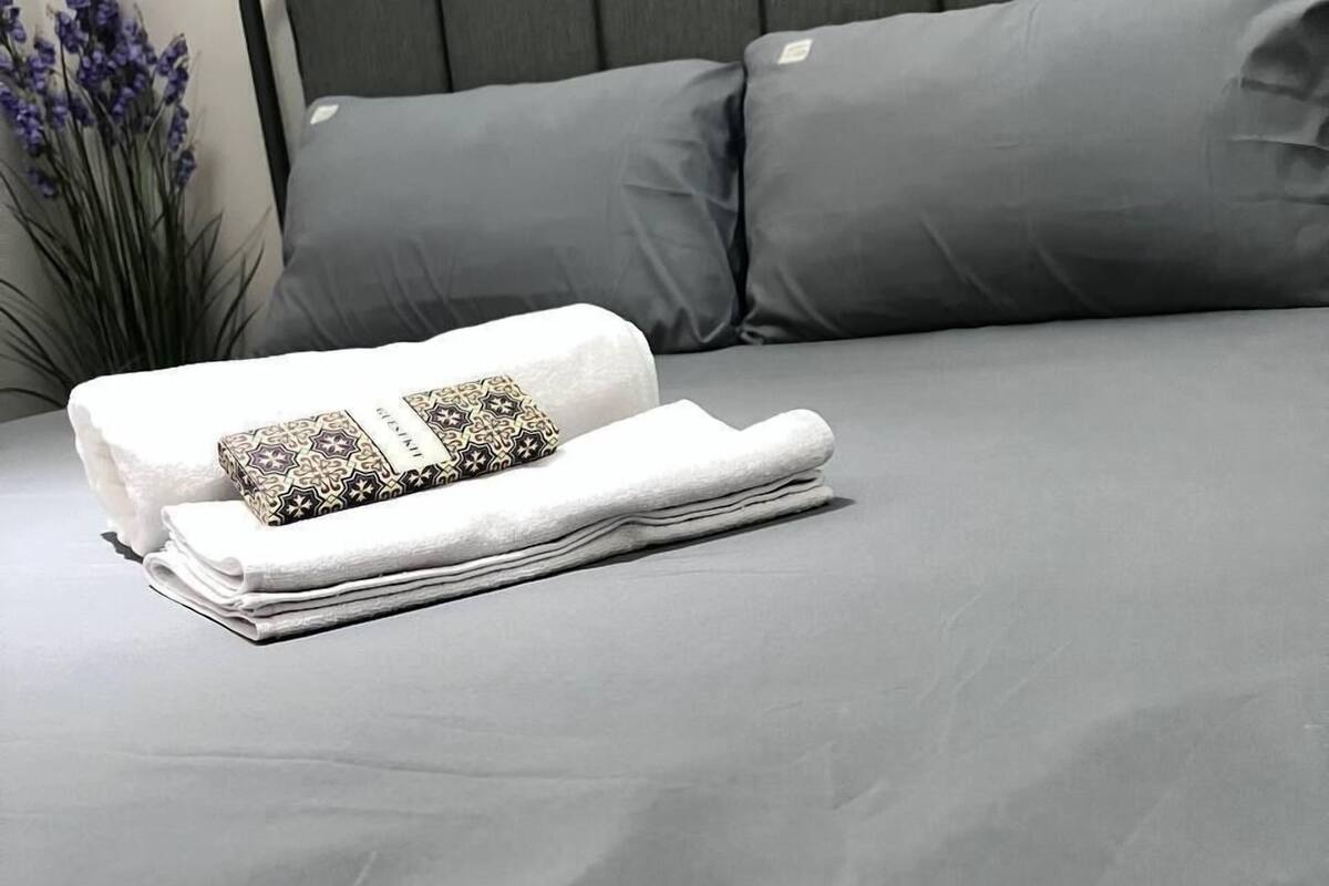 A neatly made bed with a grey mattress and pillows, a folded white towel, and a patterned pillow.