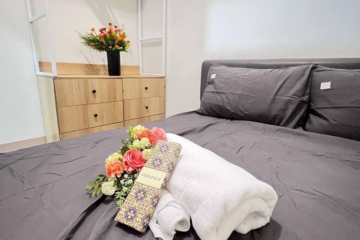 A neatly made bed with a gray bedspread, a white towel, and a floral arrangement labeled "GUEST KIT" on a hotel room.