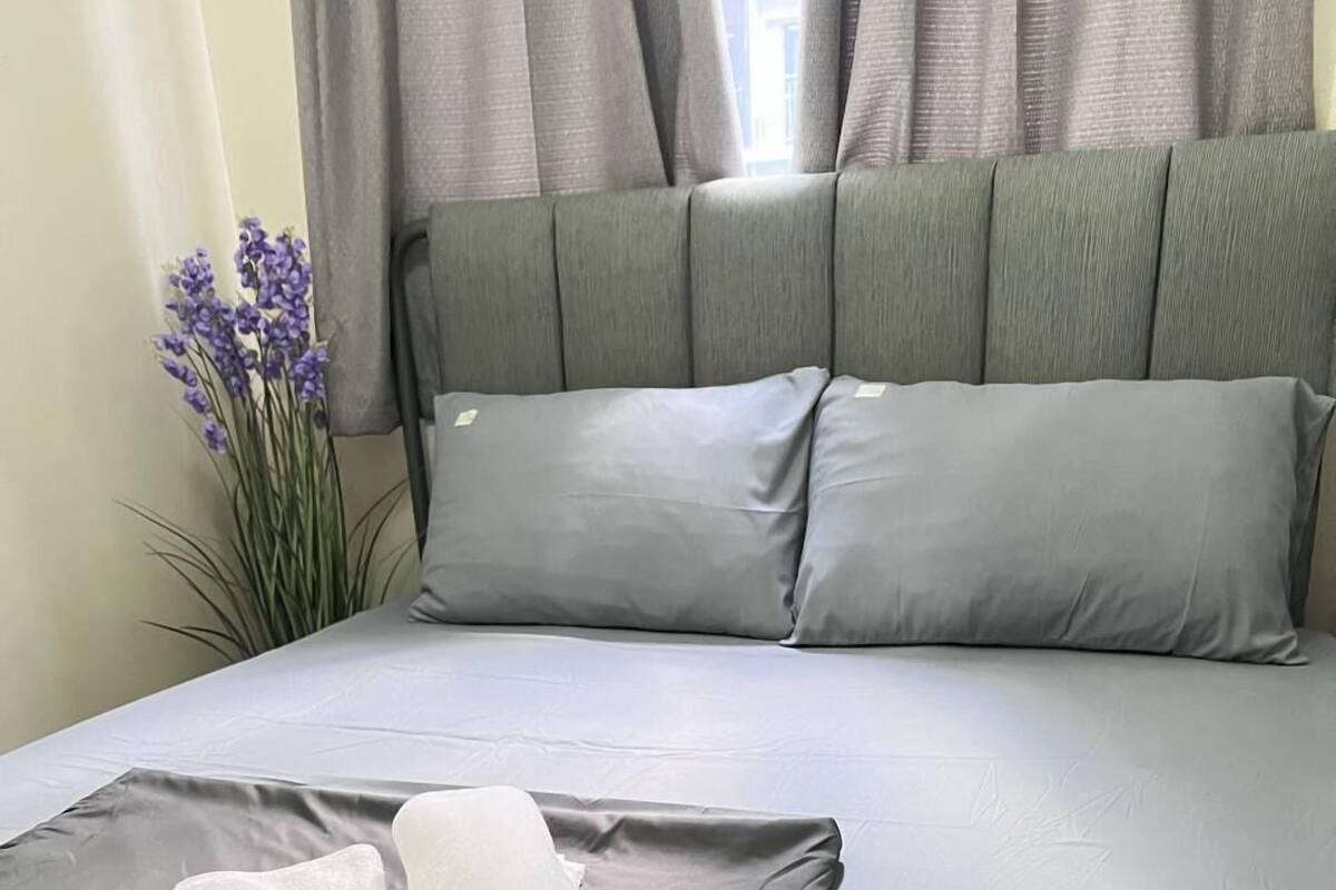 A neatly made bed with grey linens, a rolled white towel, and a folded white towel on the bed.