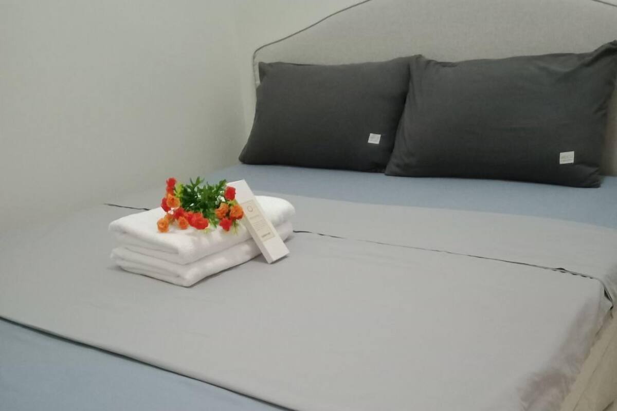 A neatly made bed with a grey headboard and pillows, a folded white towel, and a small bouquet of flowers on the bed.