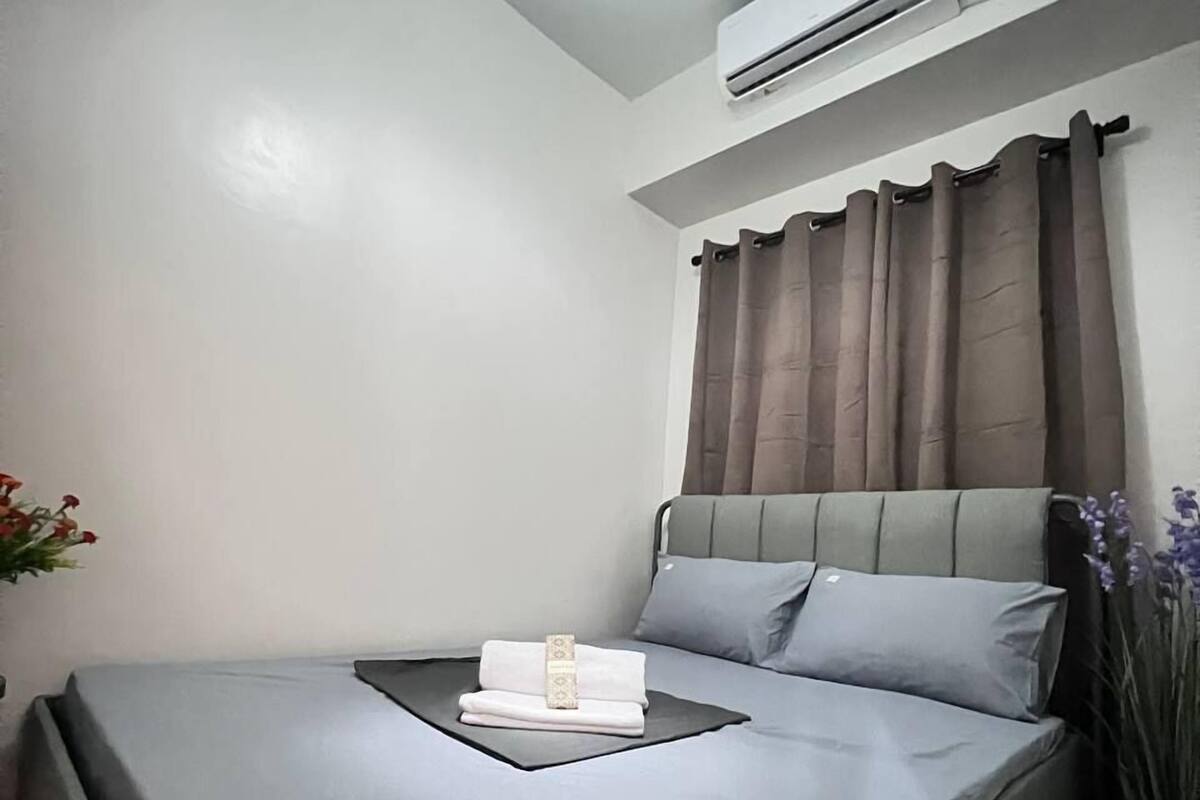 A neatly made bed with a grey mattress, a white pillow, and a folded towel. There is a ceiling light fixture, an air conditioning unit, and a curtain on the window.