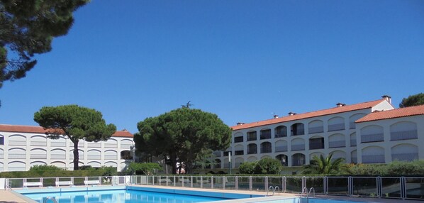 Une piscine entourée d’une clôture blanche, un grand arbre et, au fond, un bâtiment avec un toit rouge.