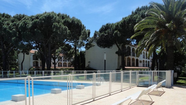 Una piscina con valla blanca, rodeada de árboles y un edificio con tejado de tejas.