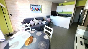 Eine moderne Wohnung mit einem grauen Sofa, einem Esstisch mit hölzerner Dekoration, einer Küche mit weißen Schränken und einer Wand in Grünton.
