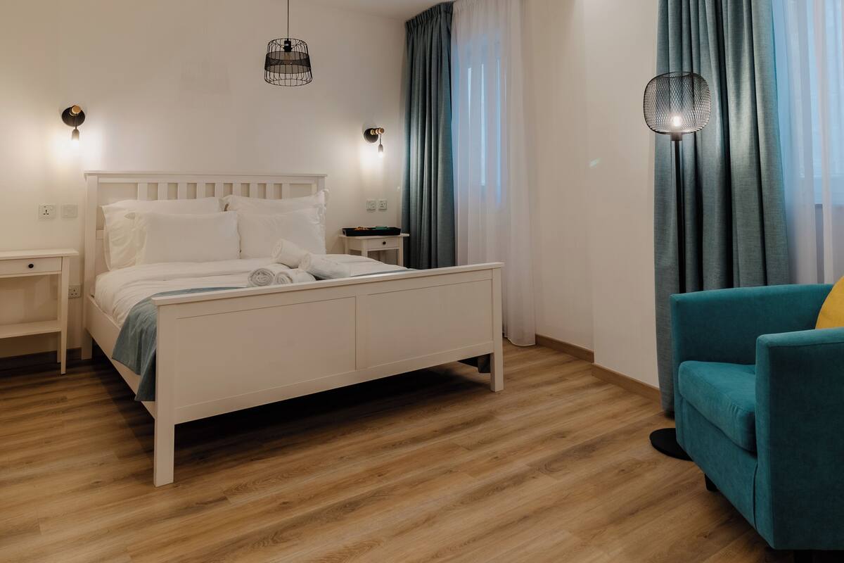 Un dormitorio con una cama blanca, dos mesitas de noche, una lámpara de pie, un sillón azul aguamarina y una ventana con cortinas.