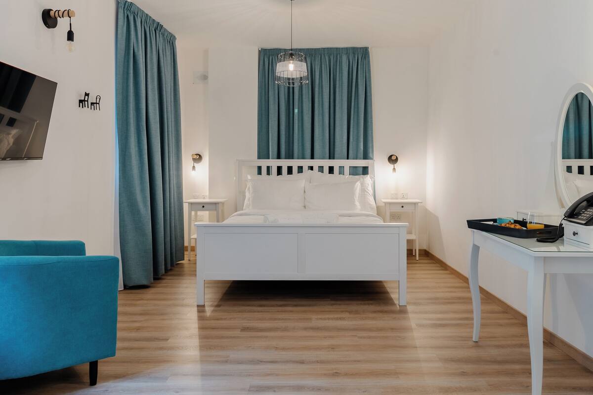 Un dormitorio moderno con una cama blanca, cortinas azul verdosas, una lámpara colgante y un escritorio con teléfono.