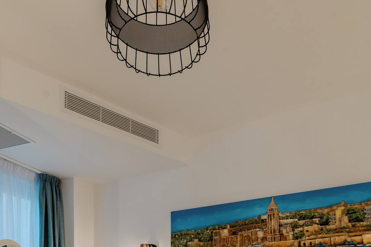 Habitación de hotel con cama, escritorio con espejo, lámpara de pared y cuadro de una ciudad costera.