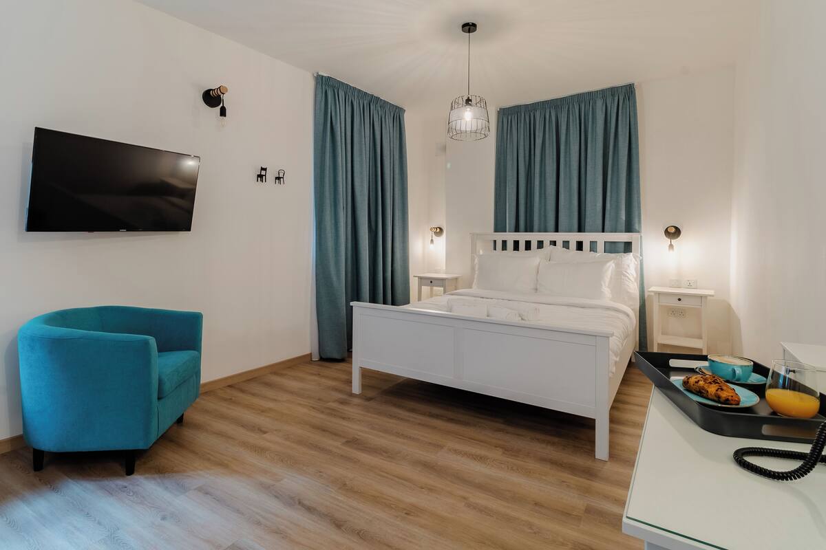 Un dormitorio moderno con una cama blanca, cortinas color turquesa, un televisor de pantalla plana, un sillón color turquesa y una mesita de noche con una bandeja de desayuno.