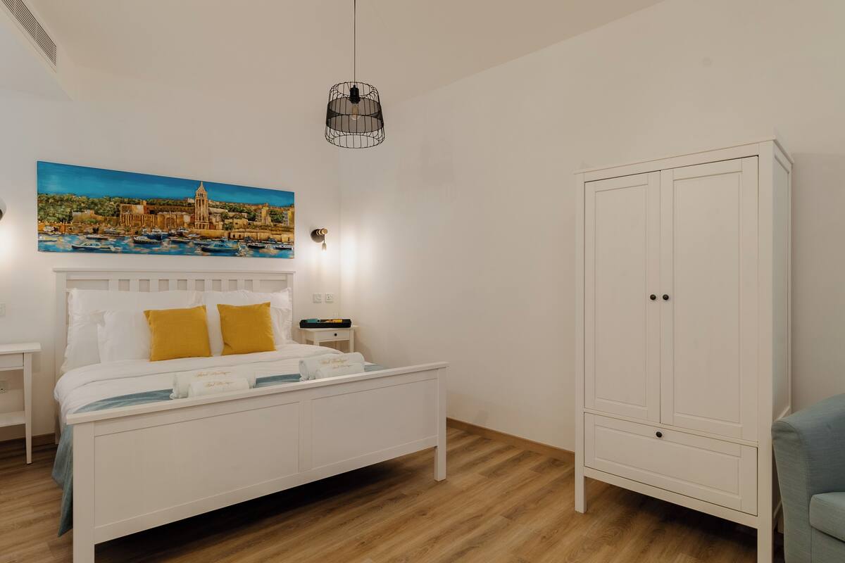 Un dormitorio con una cama, un armario blanco, una mesita de noche, un cuadro de un pueblo costero y una lámpara colgante.