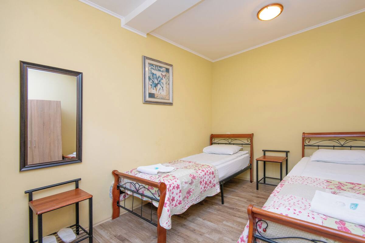 Une petite pièce propre et bien rangée, avec deux lits simples, un banc en bois, une petite table, un miroir et un tableau encadré accroché au mur.