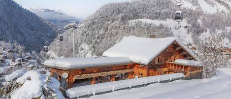 Un chalet en bois, dont le toit est recouvert de neige, entouré d’un paysage montagneux enneigé et avec un télésiège visible au loin.