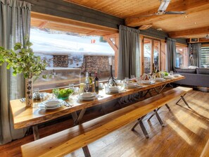 Una larga mesa de comedor de madera dispuesta para una comida, con un banco en el centro, ventanas grandes y vistas a un paisaje nevado.