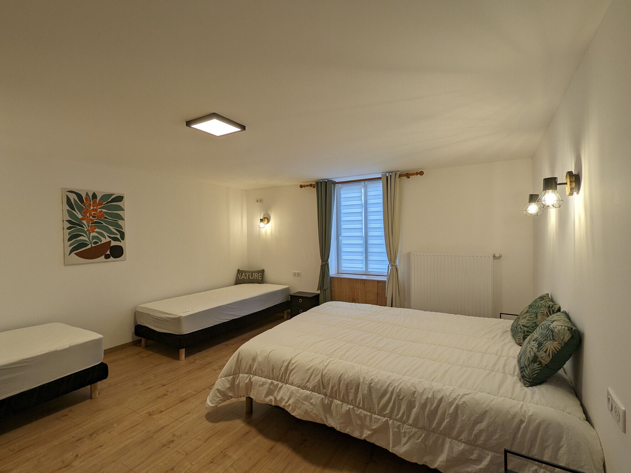 Et soveværelse med en stor seng, en mindre seng, et natbord, en lampe på væggen, et vindue med gardiner og en radiator.