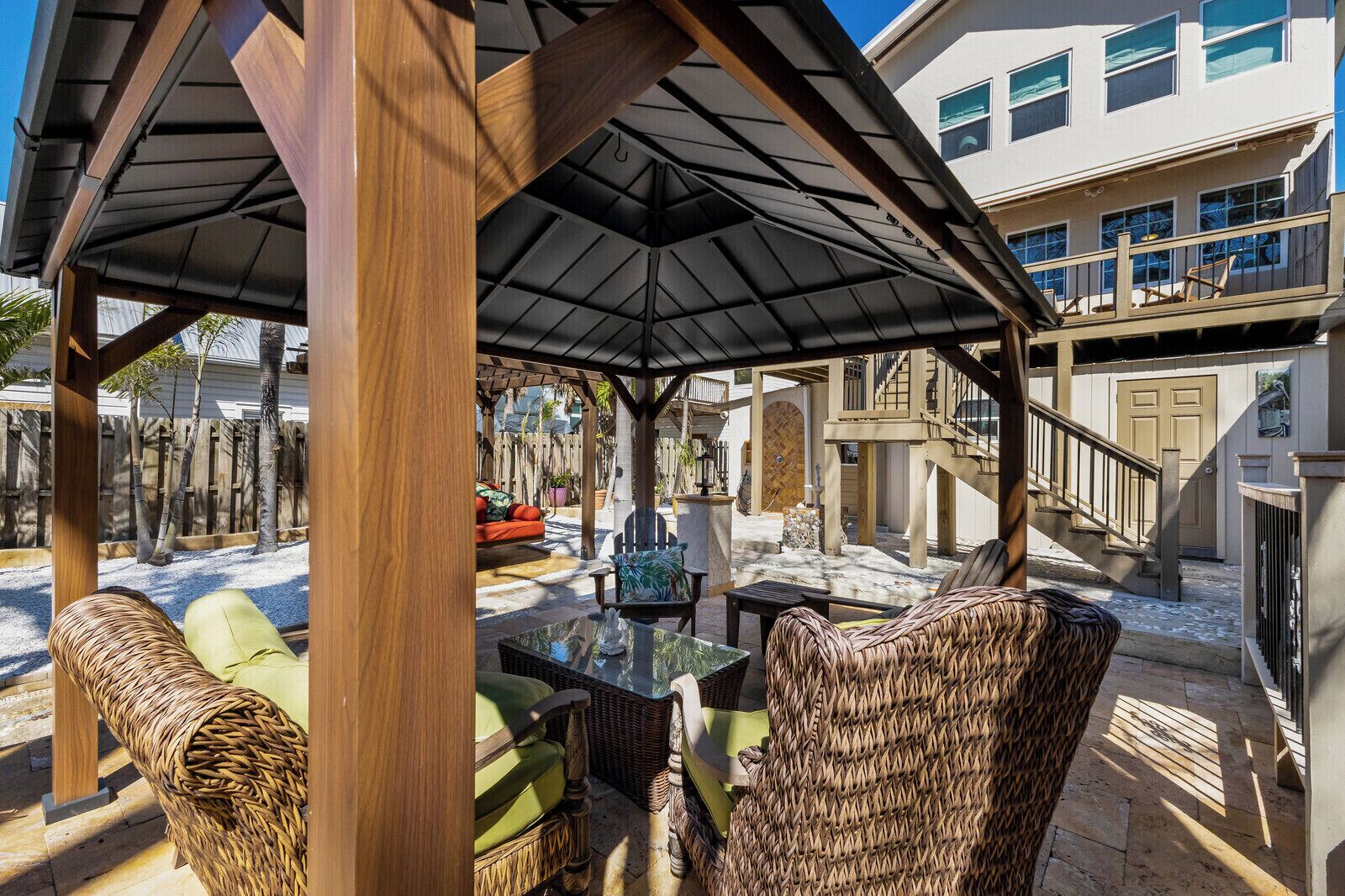 A covered outdoor seating area with wicker furniture and a central table, surrounded by a building with multiple windows and a staircase.