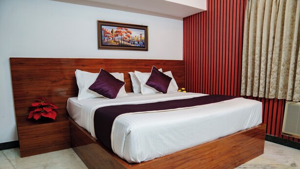 A hotel room with a wooden headboard, a large bed with white and purple bedding, a nightstand with a red flower, and a painting on the wall. - Rich Inn Palace Tnagar  (Chennai)
