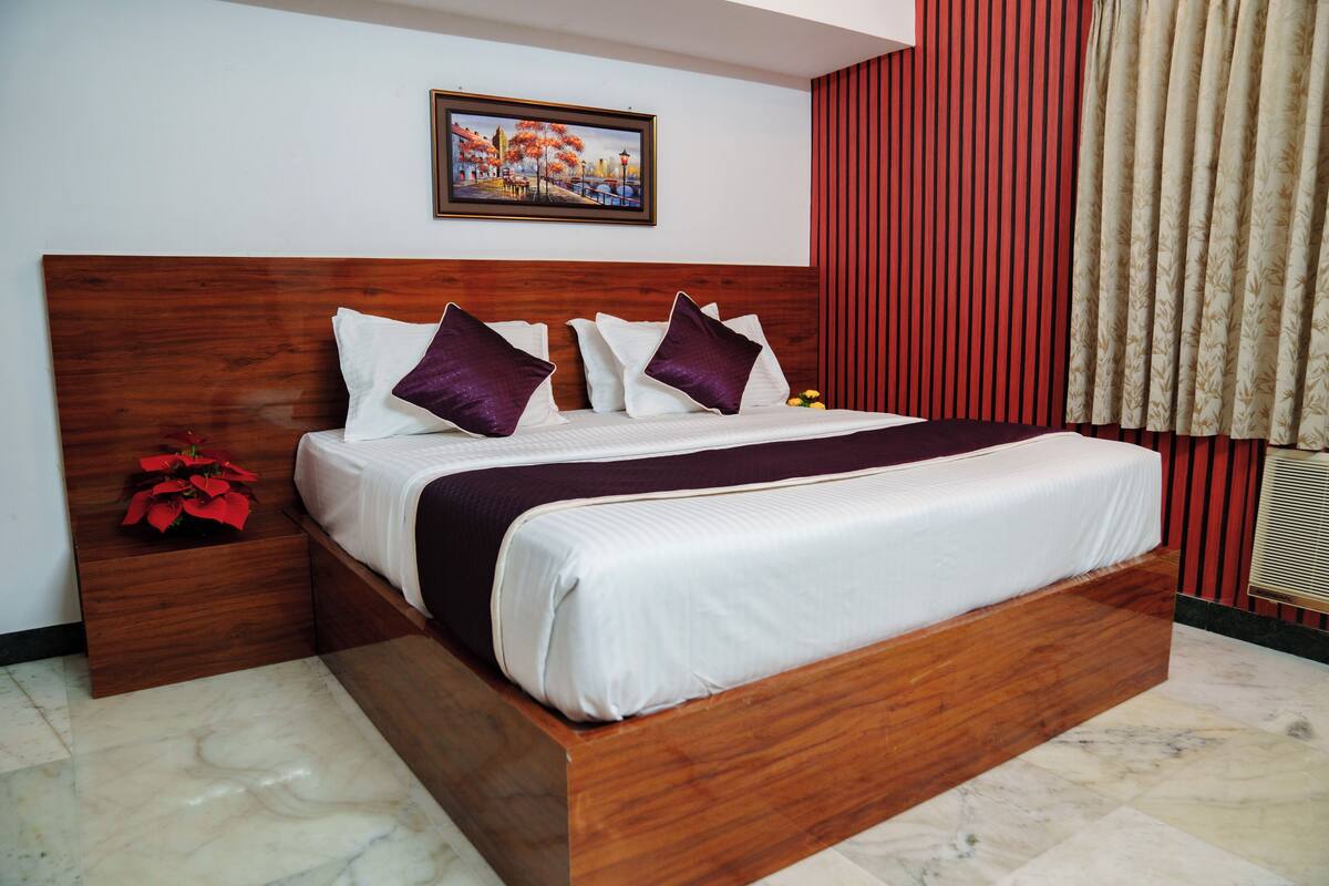 A hotel room with a wooden headboard, a large bed with white and purple bedding, a nightstand with a red flower, and a painting on the wall.