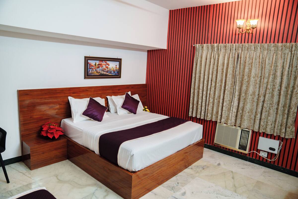 A neatly made bed with a wooden headboard, a nightstand with a red flower, and a wall-mounted air conditioning unit.