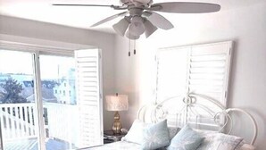 Ein Schlafzimmer mit Bett, Deckenventilator, Nachttisch, Lampe und Blick ins Freie.