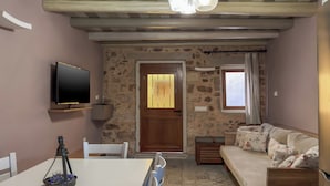 A cozy living room with a sofa, a dining table with chairs, a TV mounted on the wall, and a stone wall. - Vetus Oppidum Guesthouse (Chania)