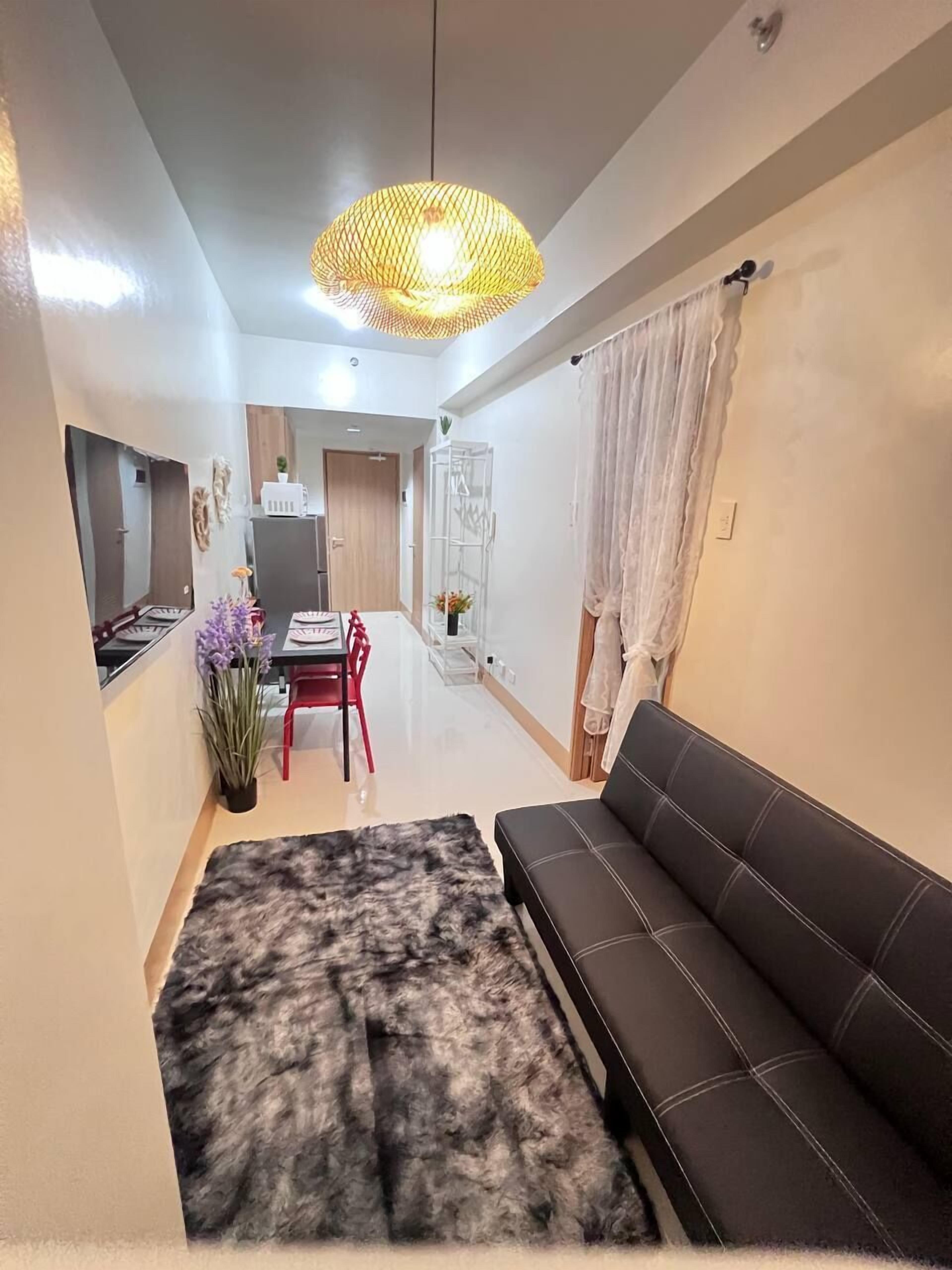 Uma sala de estar moderna com um sofá preto, um tapete estampado, uma área de jantar com mesa e cadeiras, e uma cozinha ao fundo.