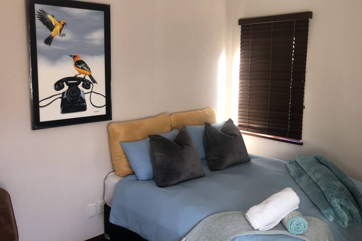 A bedroom with a bed, pillows, a blanket, a towel, a wall-mounted picture, and a window with blinds.