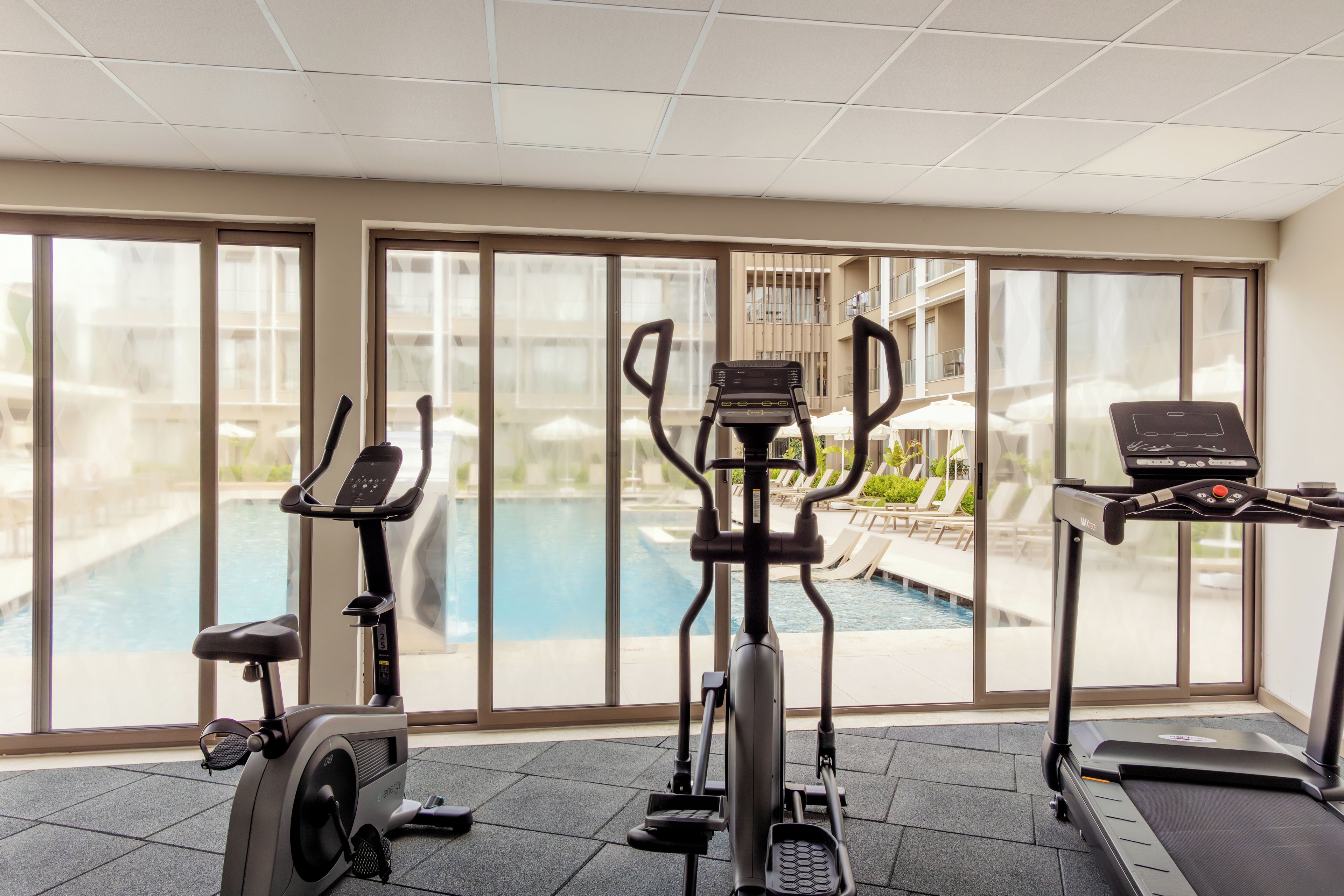 An indoor gym with exercise equipment, a pool, and a view of a building with balconies.