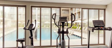 An indoor gym with exercise equipment, a pool, and a view of a building with balconies.