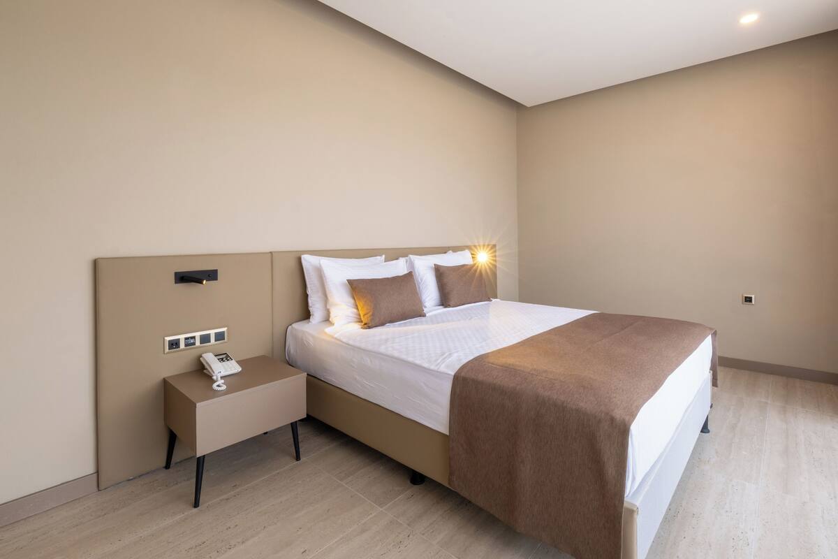 A modern hotel room with a large bed, a bedside table with a phone, and a wall-mounted light fixture.