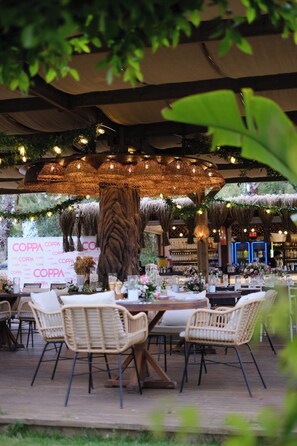 An outdoor dining area with wicker chairs, tables set with white tablecloths, and a large tree trunk centerpiece.