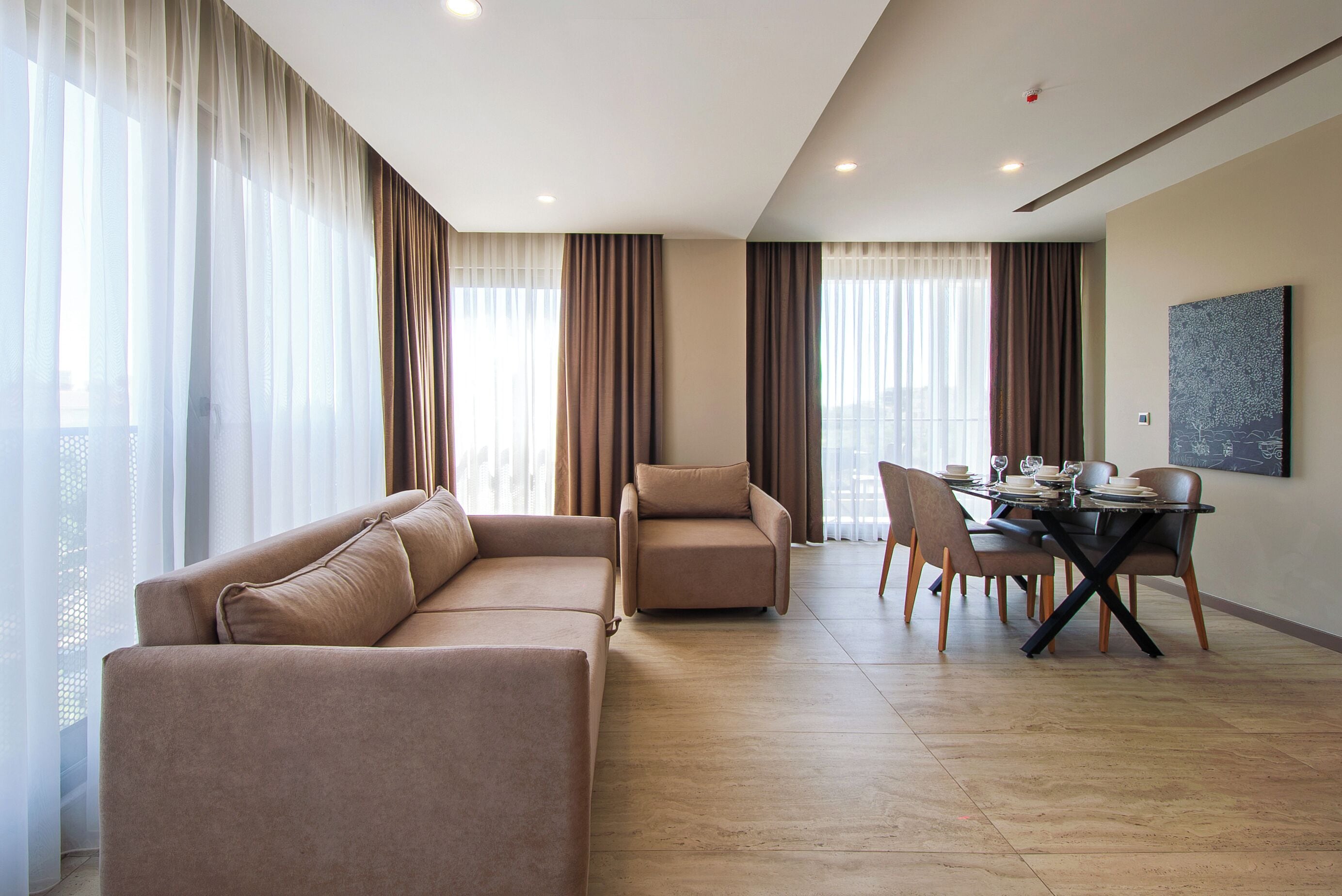 Een moderne woonkamer met een hoekbank, een fauteuil, een eettafel met stoelen, en een groot raam met dunne gordijnen.