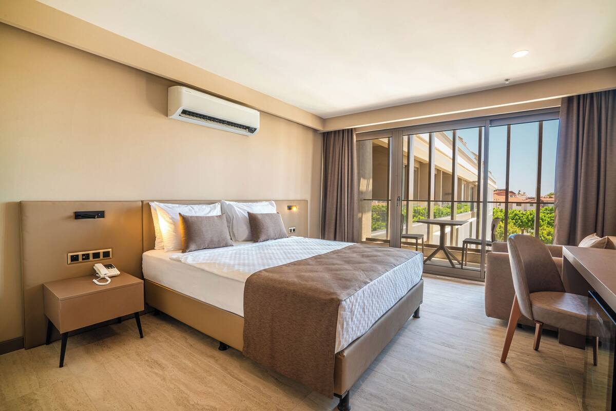 A modern hotel room with a large bed, bedside table with a phone, and a view of the outdoors through a sliding glass door.