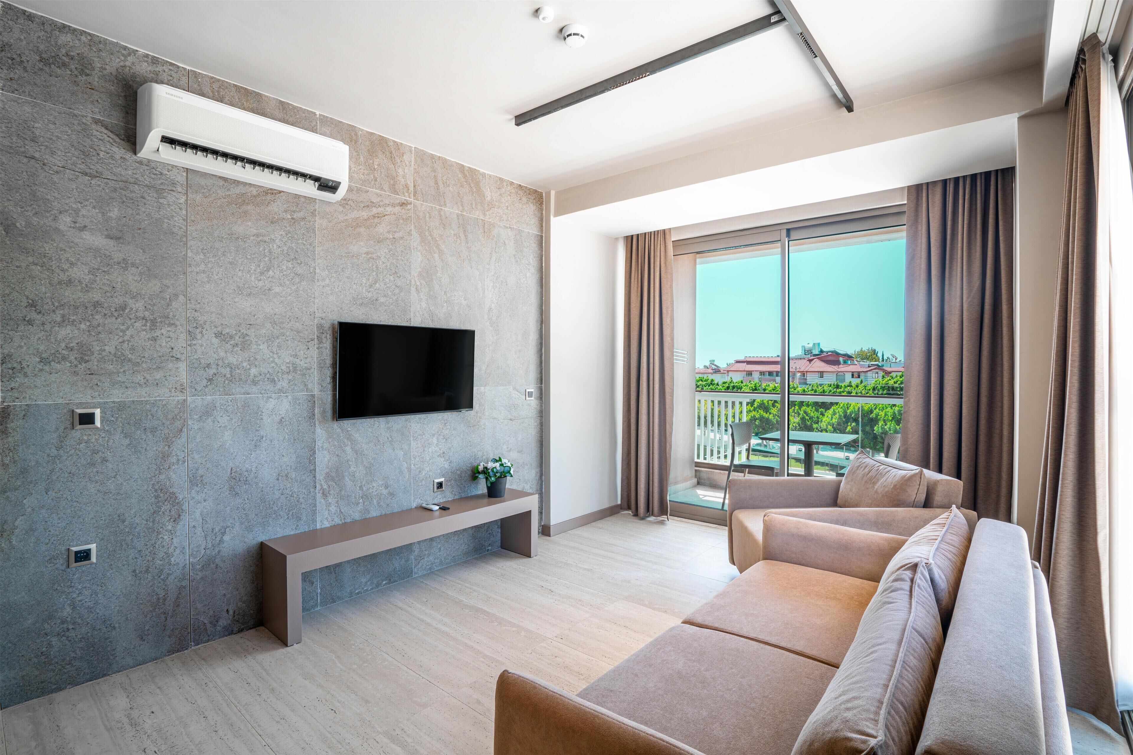 A modern living room with a flat-screen TV mounted on the wall, a large window with curtains, a beige sofa, and a bench.
